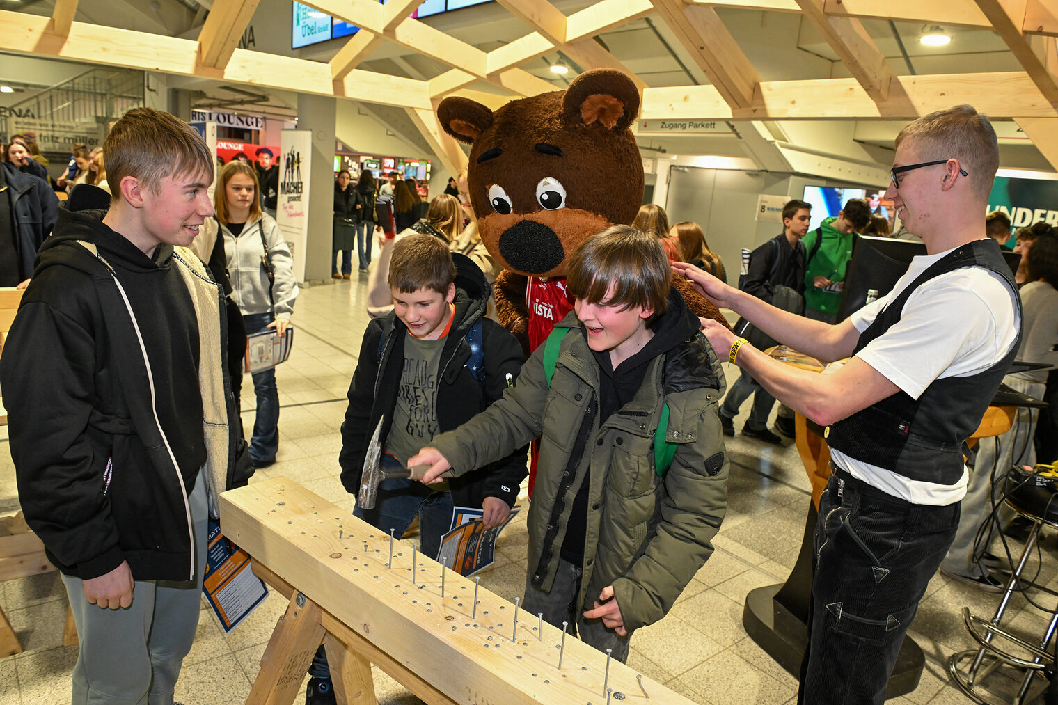 Zwei Schüler hämmern Nägel in einen Holzbalken während Maskottchen Freaky zusieht.
