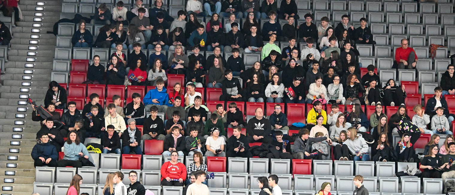 Die Schülerinnen und Schüler sitzen auf der Nordtribüne und sehen sich das Spiel an.
