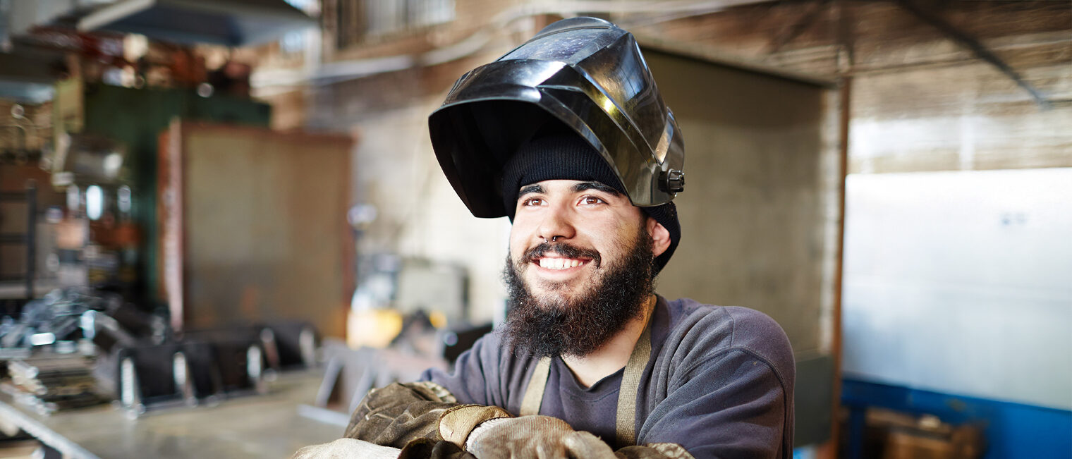 Ein Schweißer trägt einen Schutzhelm in der Werkstatt und lächelt in die Kamera.
