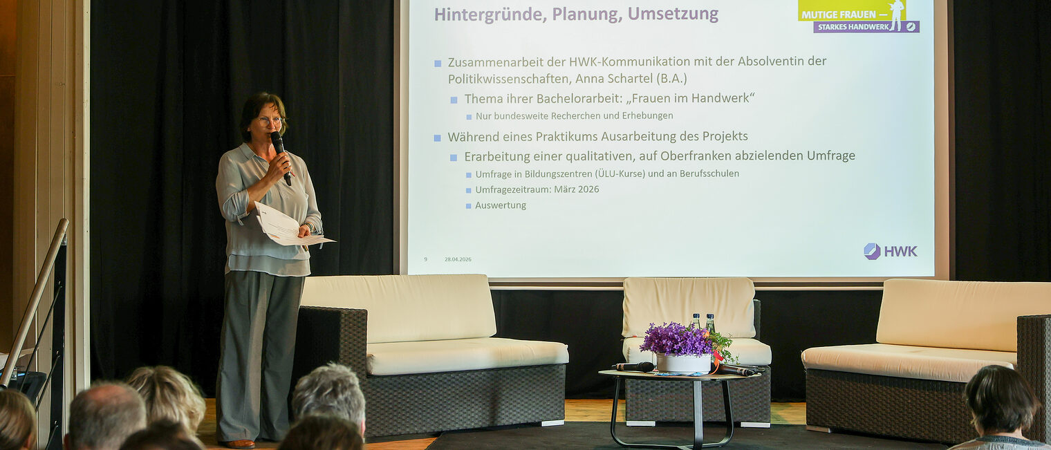 Michaela Heimpel steht vor einer Leinwand auf dem Podium und spricht in ein Mikrofon.