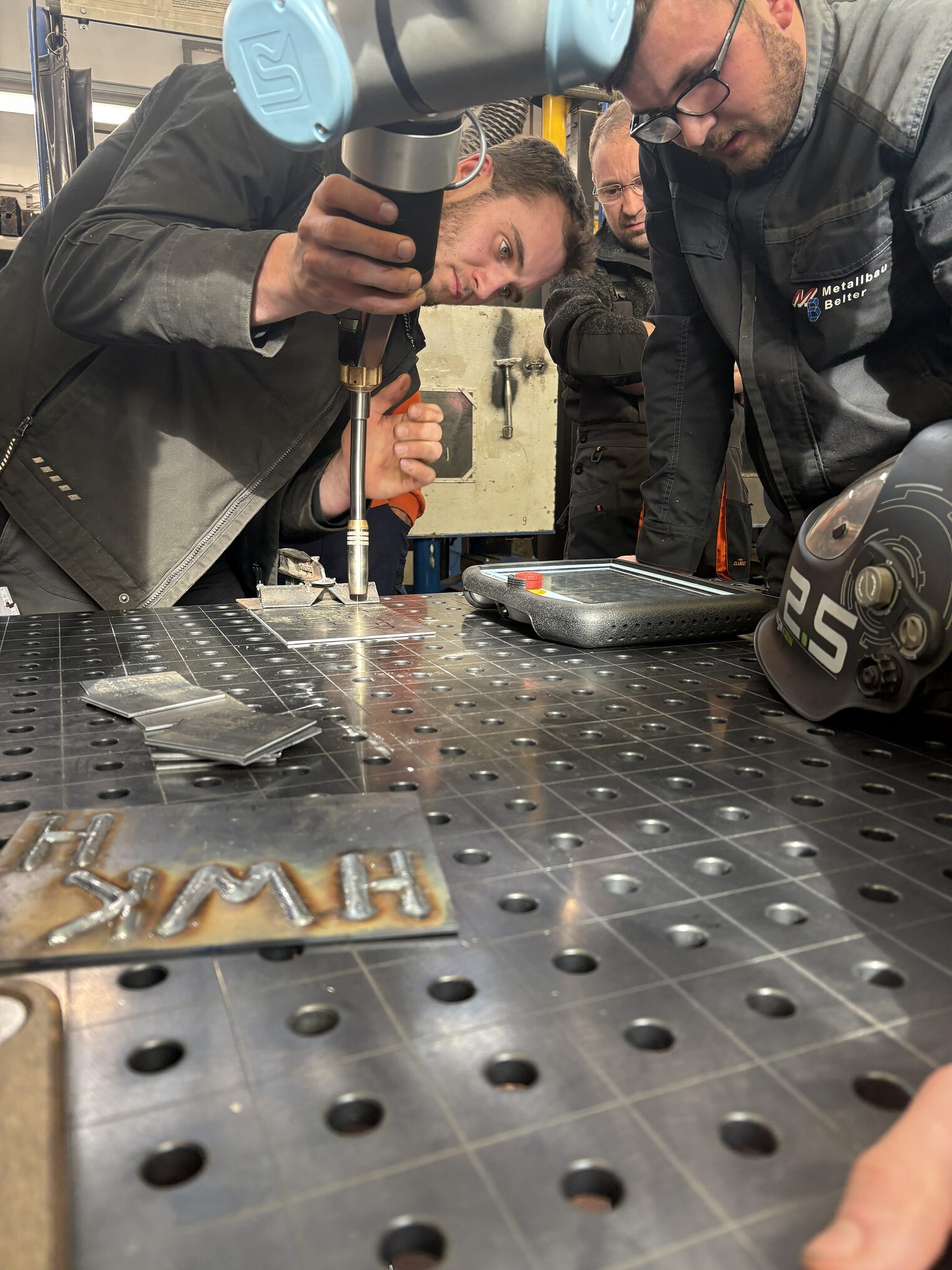 Eine Gruppe von Meisterschülern nutzt einen Schweißroboter, um Schrift in Metallteile zu gravieren.