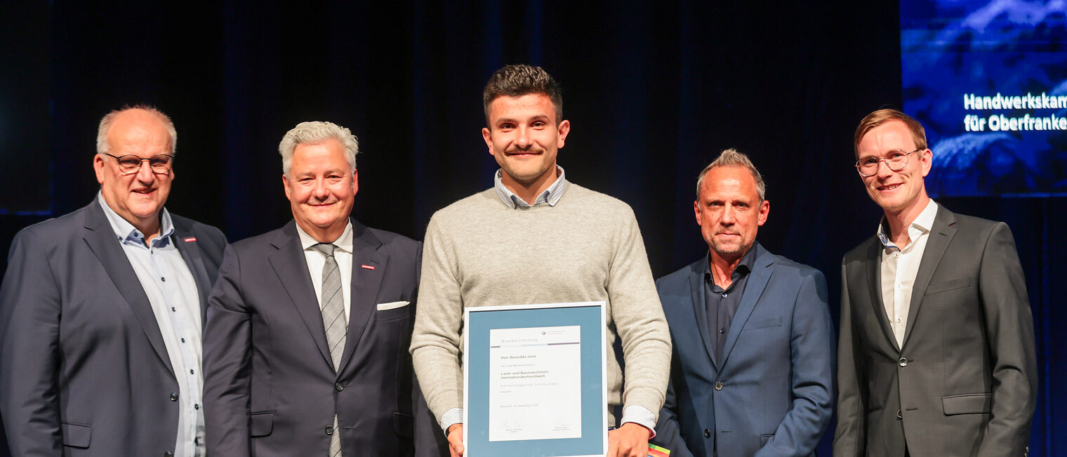 Jungmeister Benedikt Joos präsentiert seine Ehrenurkunde mit Gratulanten auf der Bühne.