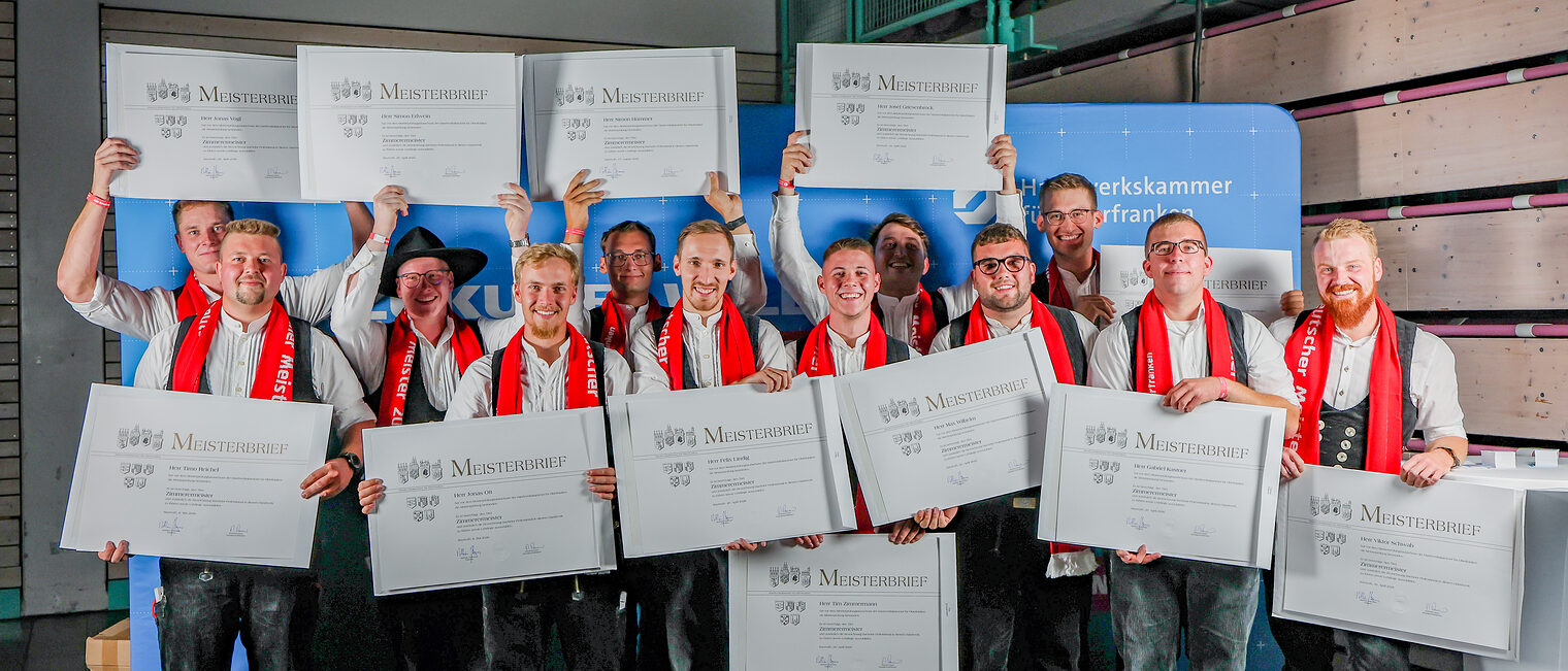 Die Jungmeister zum Zimmerermeister posieren für ein Gruppenfoto vor einer Fotowand.