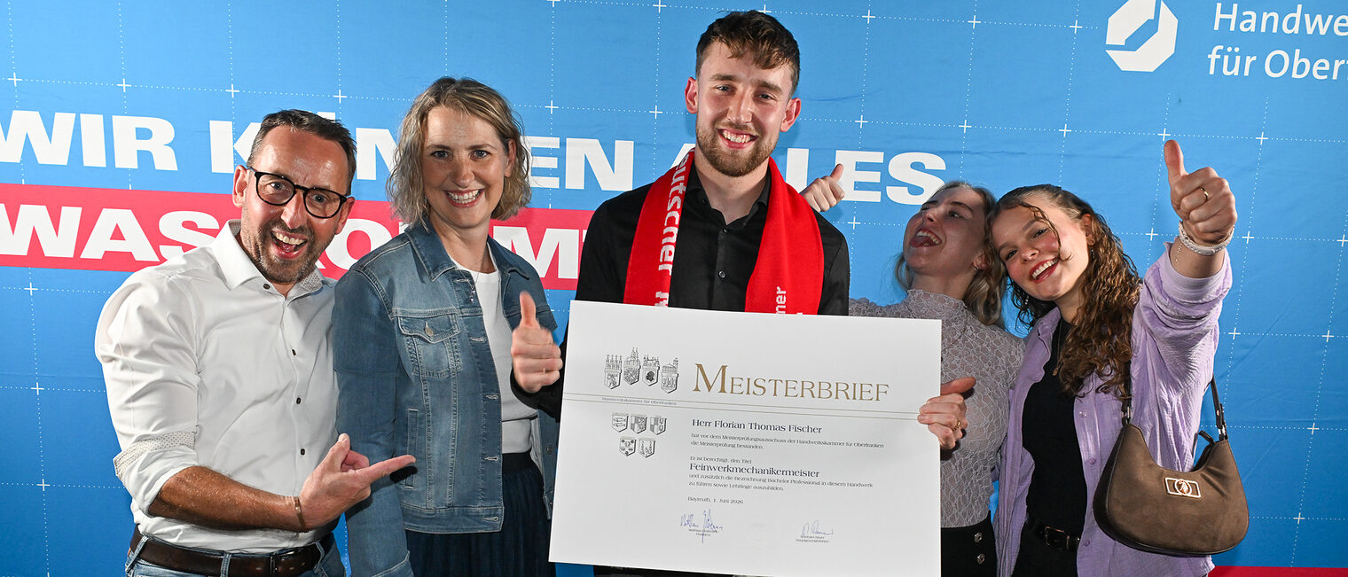 Jungmeister Florian Thomas Fischer posiert mit seinem Meisterbrief und seiner Familie vor einer Fotowand.