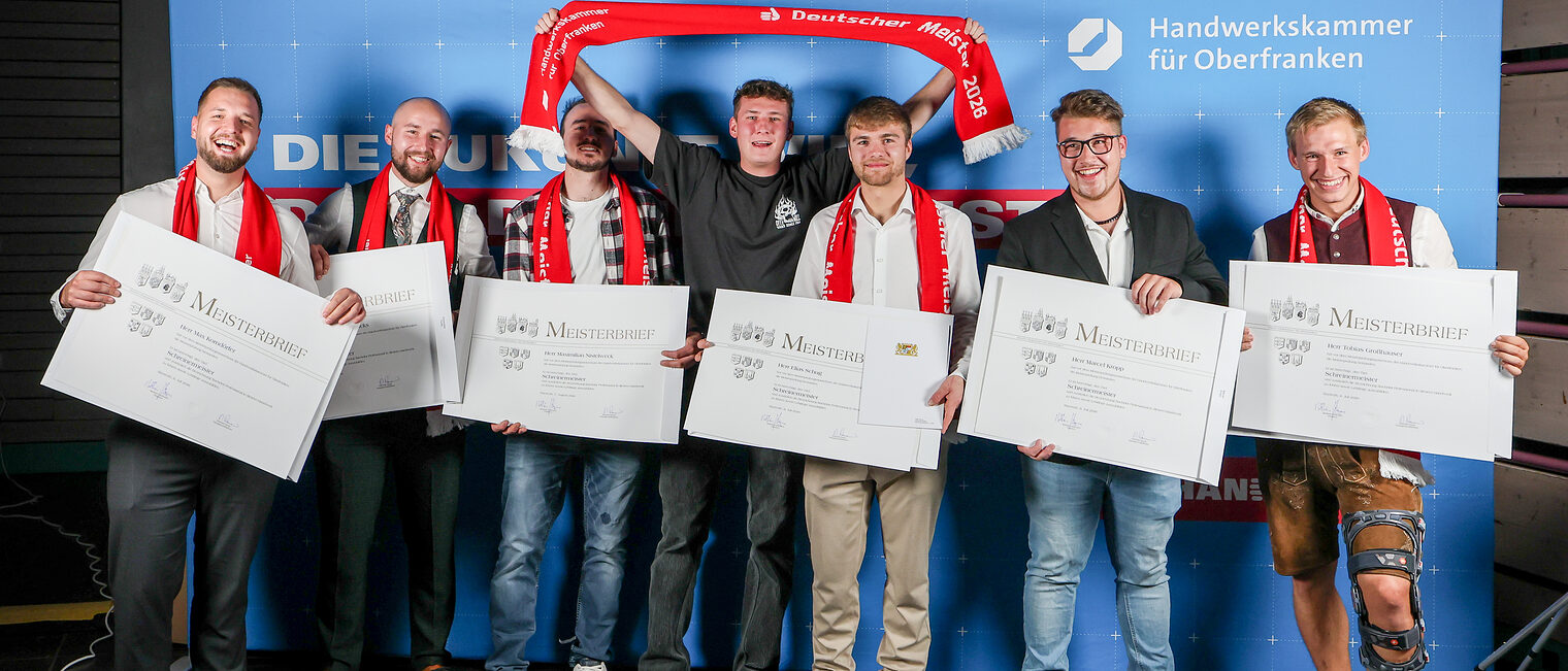 Eine Gruppe Meister präsentiert ihre Meisterbriefe vor einer Fotowand.