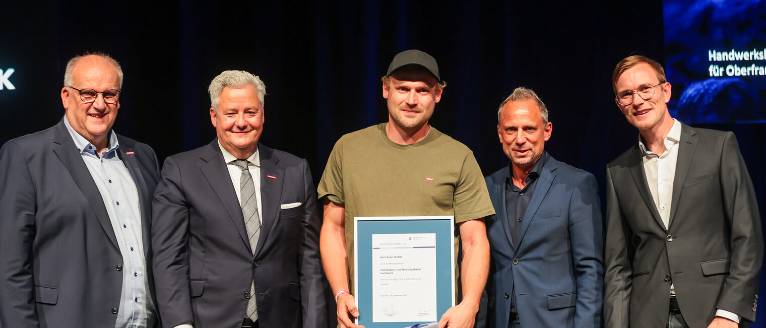 Der Prüfungsbeste Sven Distler zeigt seine Urkunde mit Gratulanten auf der Bühne.