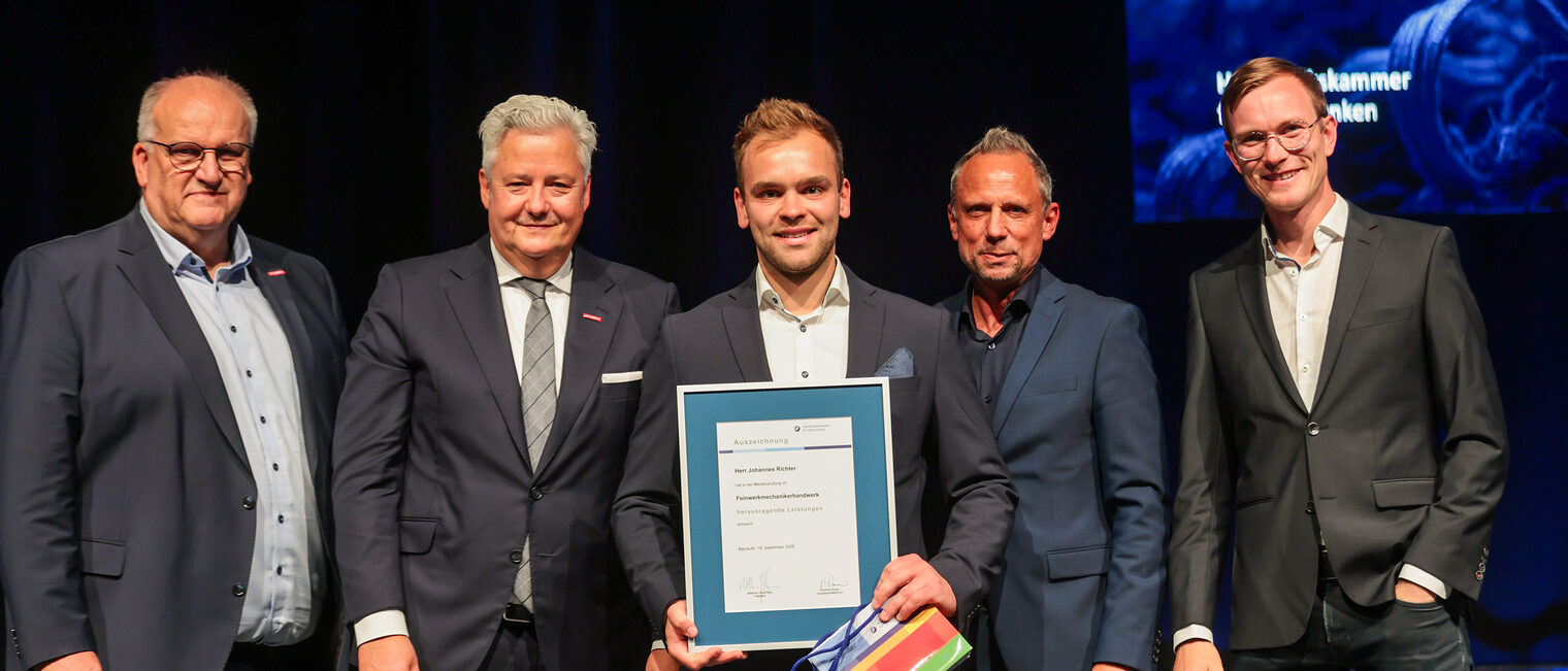 Der Prüfungsbeste Johannes Richter zeigt seine Urkunde mit Ehrengästen auf der Bühne.