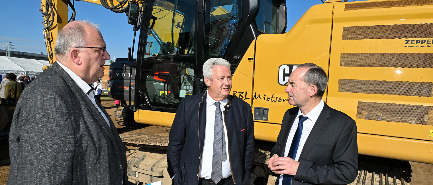 Hubert Aiwanger redet vor einer Baumaschine mit Reinhard Bauer und Matthias Graßmann.