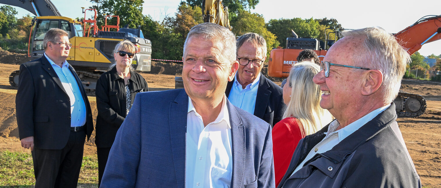 Gäste der Baustartfeier unterhalten sich vor Baggern.