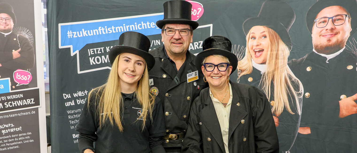 Mitarbeiter eines Betriebs aus dem Schornsteinfegerhandwerk stehen in Arbeitskleidung und mit Hut zusammen.
