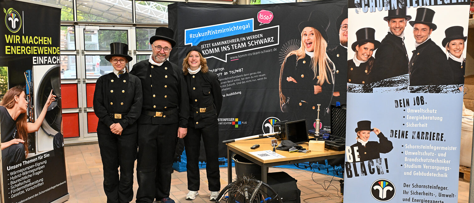 Das Team eines Kaminkehrerbetriebes steht in Arbeitskleidung mit Hut an seinem Stand.