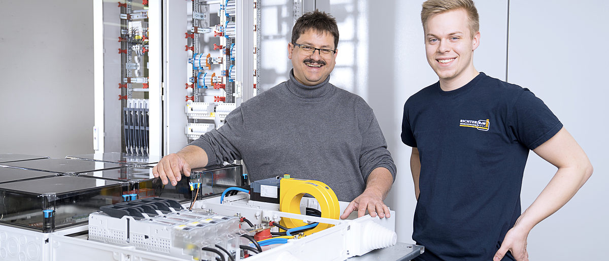 Bernd Zeilmann (im Bild links), Geschäftsführer der Richter R&W Steuerungstechnik GmbH in Ahorntal, und Sebastian Regus, Ausbildungsmeister und Montageleiter, präsentieren Komponenten für den intelligenten Energieknoten (IEK). Für das Baukastensysten stehen verschiedene Module zur Verfügung, unter anderem ein iMSys- und Einspeisemodul (iMSys: intelligentes Messmodul, im Bildvordergrund liegend) und ein Niederspannnungsverteilermodul (im Bildhintergrund stehend). Bernd Zeilmann (im Bild links), Geschäftsführer der Richter R&W Steuerungstechnik GmbH in Ahorntal, und Sebastian Regus, Ausbildungsmeister und Montageleiter, präsentieren Komponenten für den intelligenten Energieknoten (IEK). Für das Baukastensysten stehen verschiedene Module zur Verfügung, unter anderem ein iMSys- und Einspeisemodul (iMSys: intelligentes Messmodul, im Bildvordergrund liegend) und ein Niederspannnungsverteilermodul (im Bildhintergrund stehend). Schlagwort(e): Energiewende, IeK, Intelligenter Energieknoten, DC-Netze, Gleichstrom, iSyme, intelligente Symmetrieeinrichtung, iMSys, Modulare Steuereinheit, DC-Grid-Manager, Seifriz-Preis, R&W Steuerungstechnik, Universität Bayreuth, HWK Oberfranken, Fraunhofer IISB